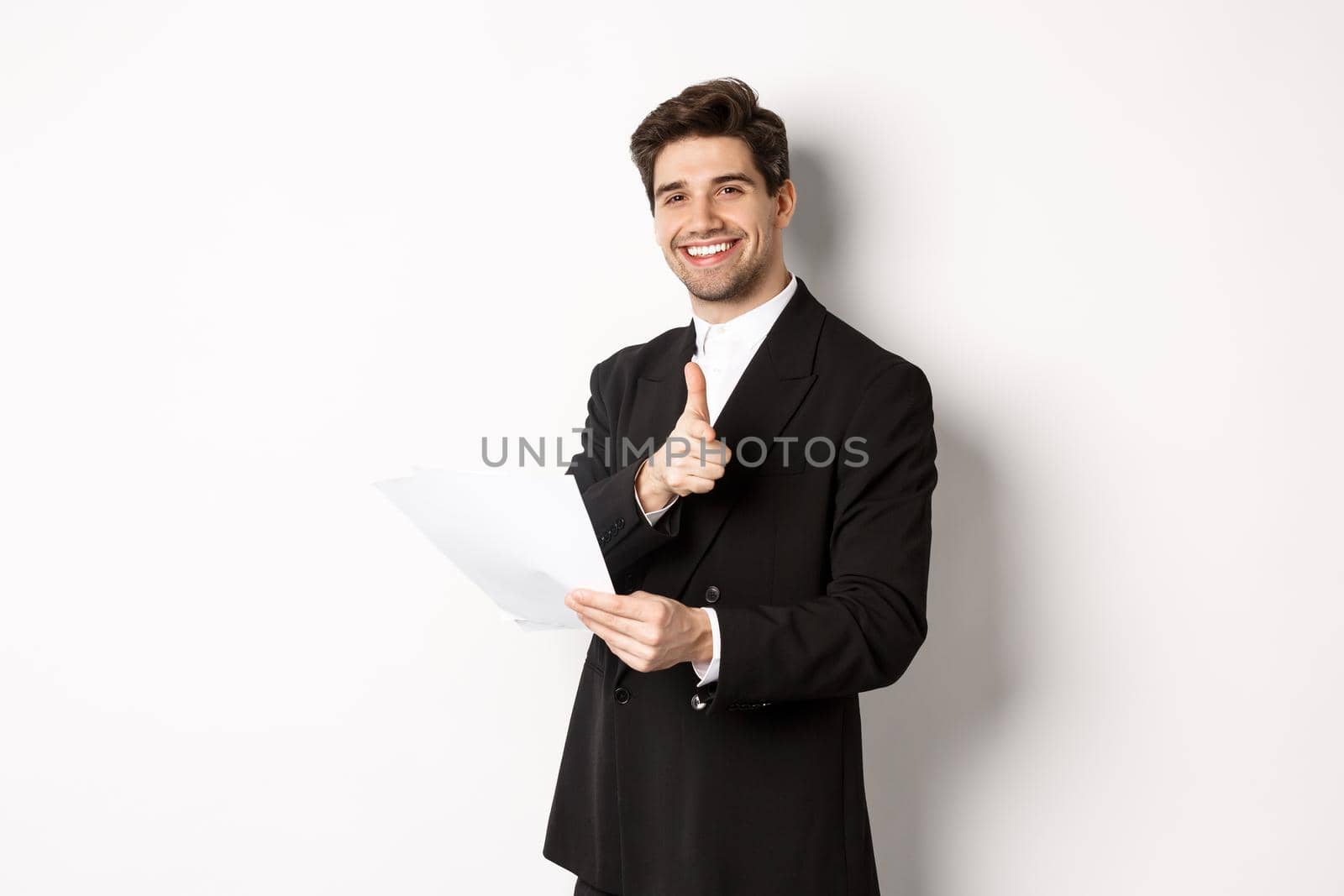 Image of handsome businessman in black suit, holding document and pointing finger at camera, praising good job, standing against white background by Benzoix