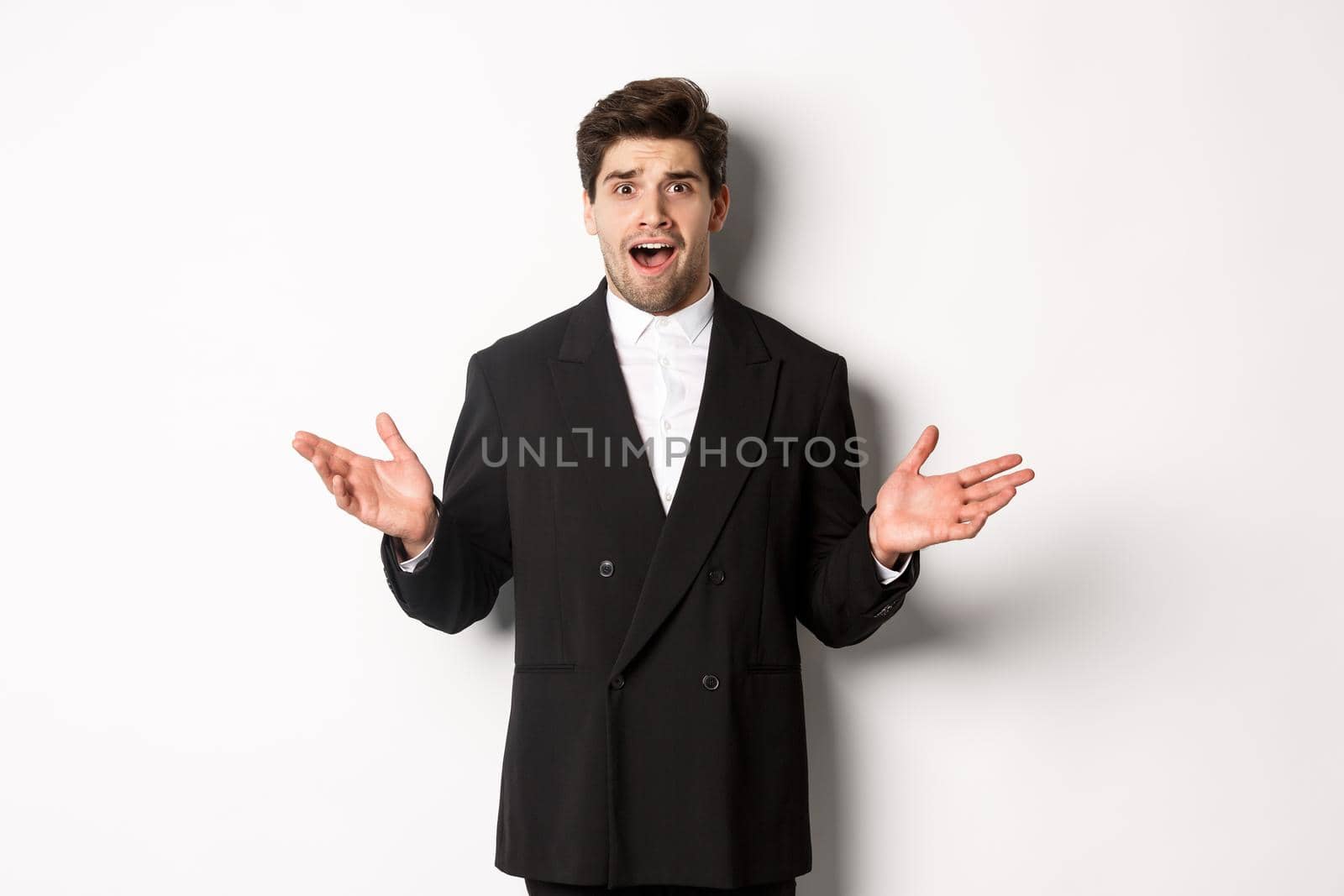 Portrait of confused and worried handsome man in suit, looking at something strange, spread hands sideways and standing puzzled against white background.