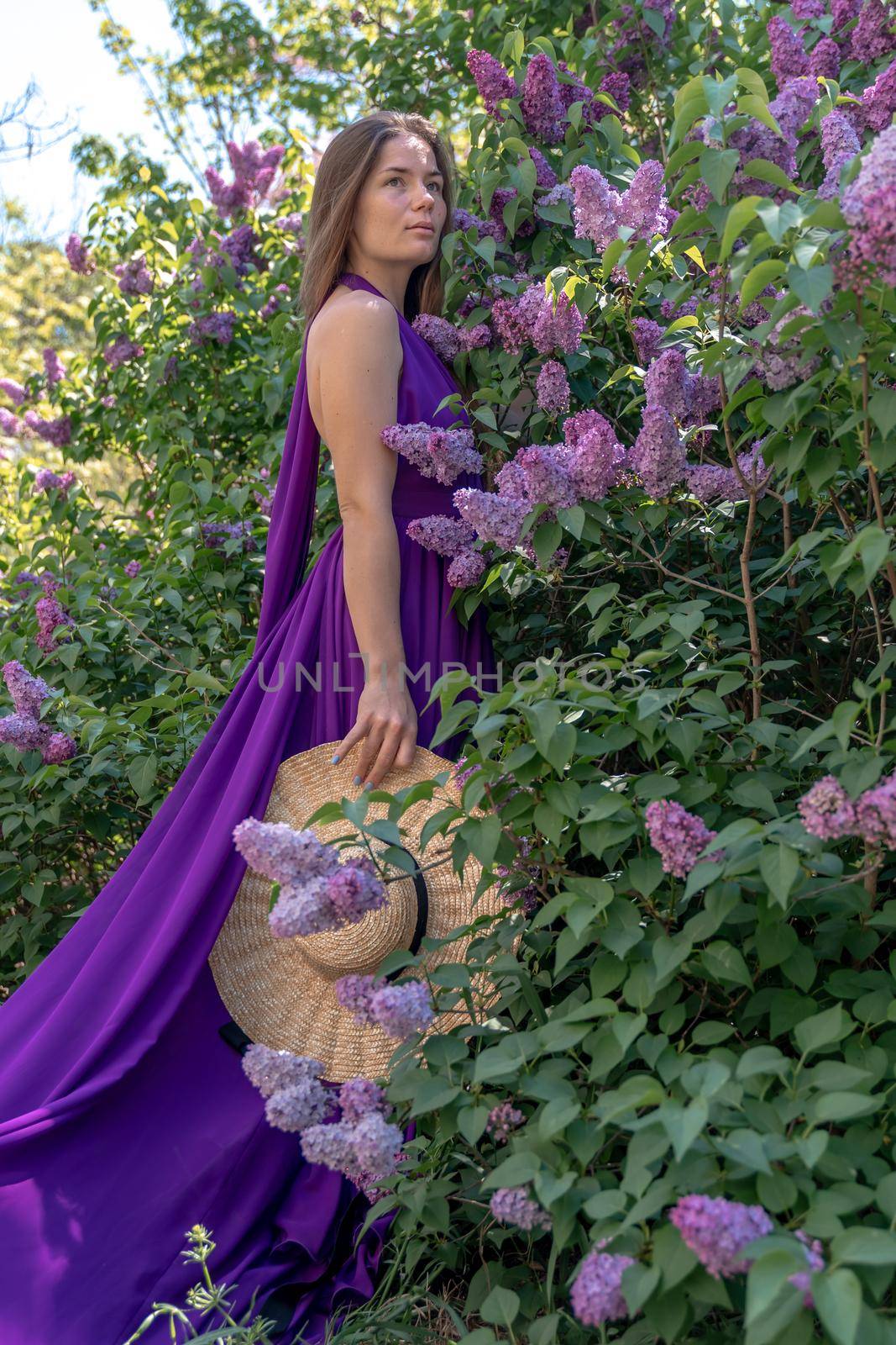 Fashion Model in Lilac Flowers, Young Woman in Beautiful Long Dress Waving on Wind, Outdoor Beauty Portrait in Blooming Garden.