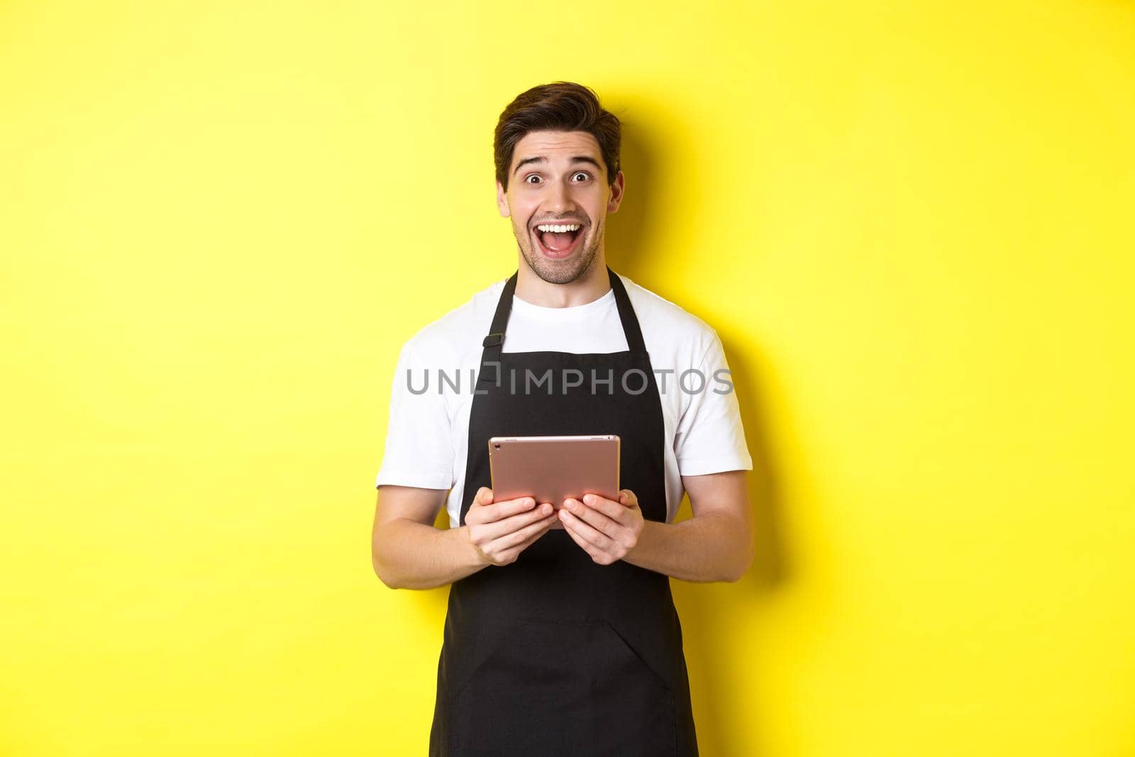 Happy salesman in black apron, holding digital tablet and looking surprised, standing against yellow background by Benzoix