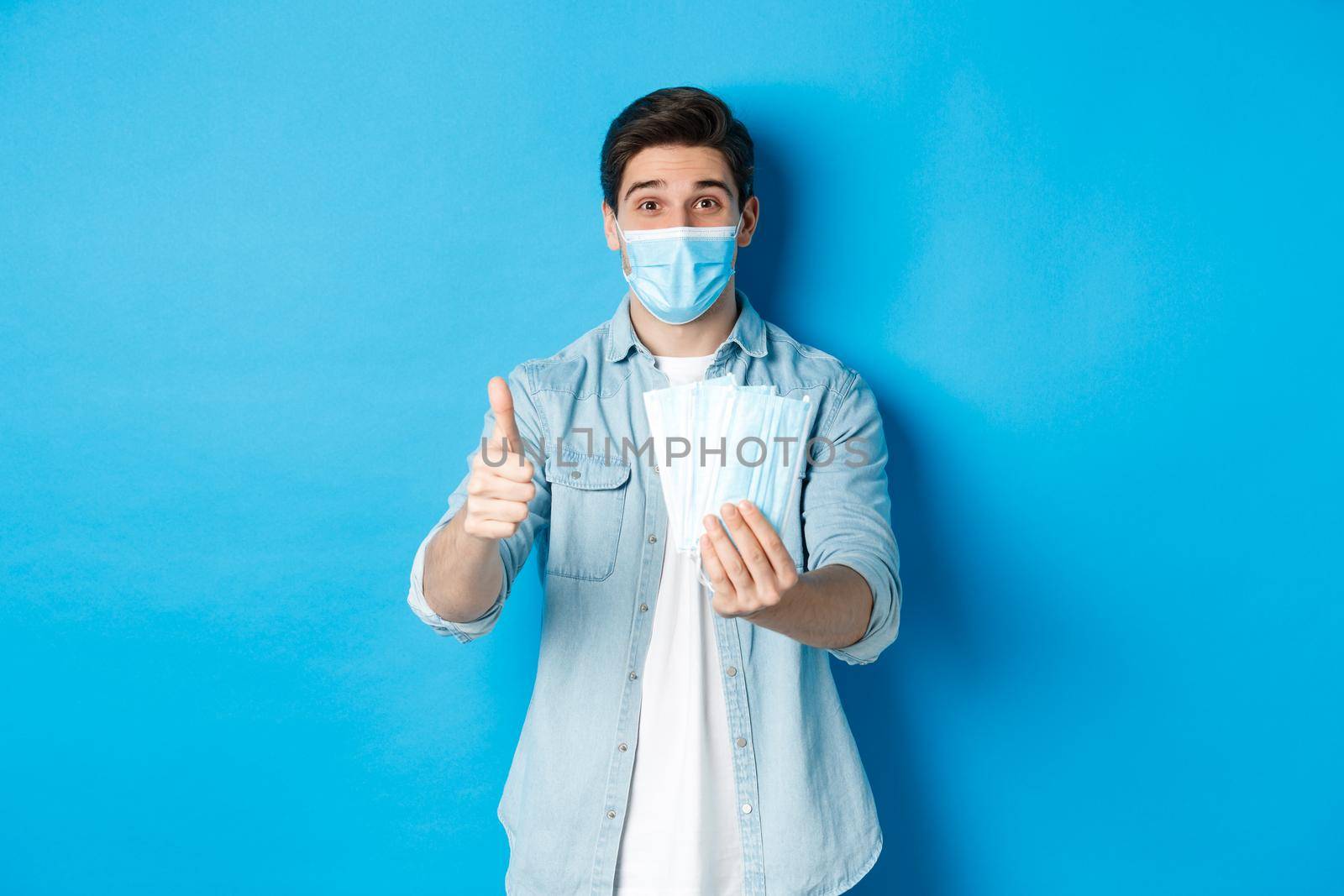 Concept of coronavirus, quarantine and social distancing. Young man showing medical masks and thumb up, standing against blue background by Benzoix