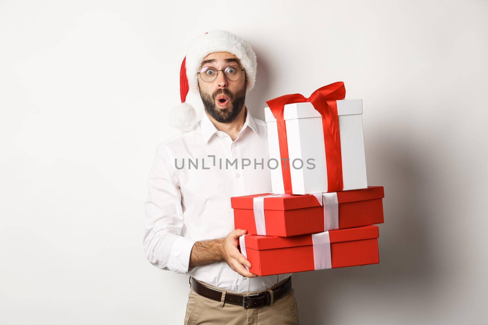 Winter holidays and celebration. Excited man holding Christmas gifts and looking surprised, wearing Santa hat, standing over white background.