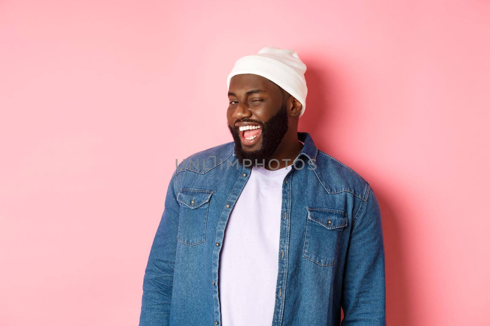 Cheeky african-american man winking at camera and smiling, teasing with offer, standing in hipster clothing against pink background.