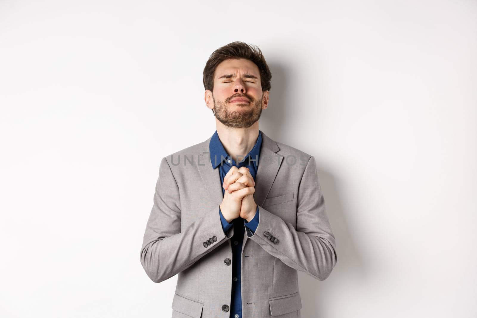 Nervous male entrepreneur in suit begging god, holding hands in pray with pleading expression, close eyes and make wish, asking for help, standing on white background by Benzoix