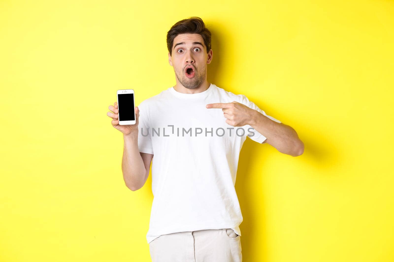 Impressed guy check out smartphone screen, pointing finger at display and gasping amazed, standing over yellow background. Copy space