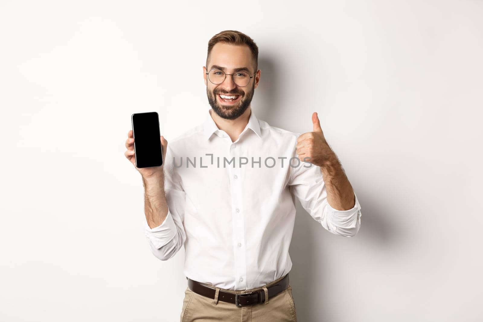 Satisfied business man in glasses showing thumbs up and demonstrating mobile phone screen, recommending app, standing over white background by Benzoix