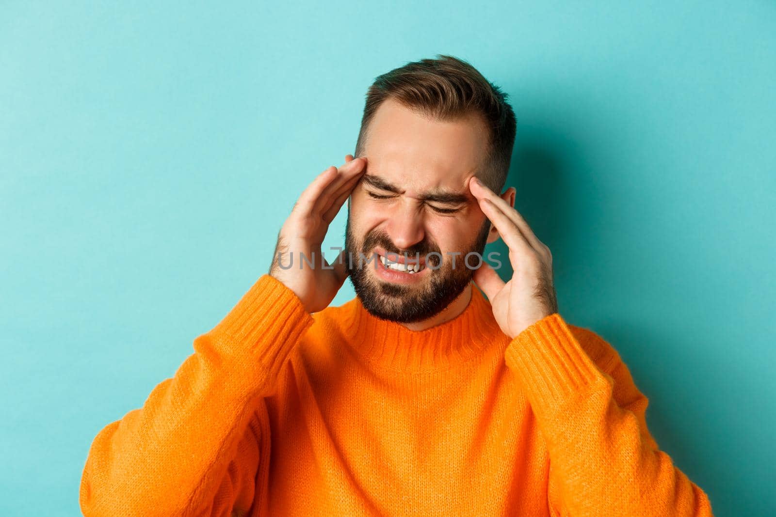 Close-up of troubled man having headache, grimacing and touching head, suffering migraine, standing over light blue background.