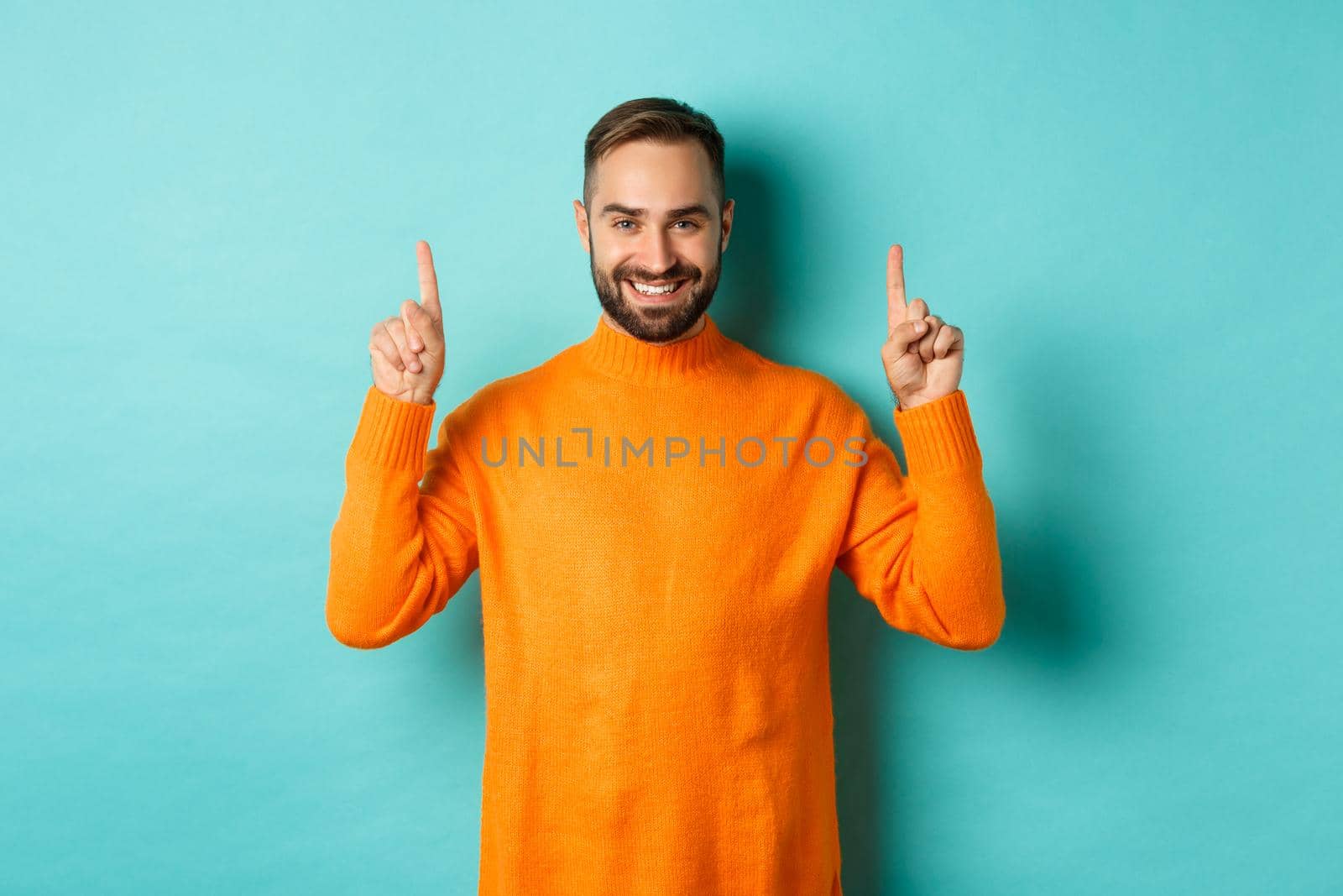 Handsome bearded guy in orange sweater showing up promo, pointing at top and smiling, standing over turquoise background by Benzoix