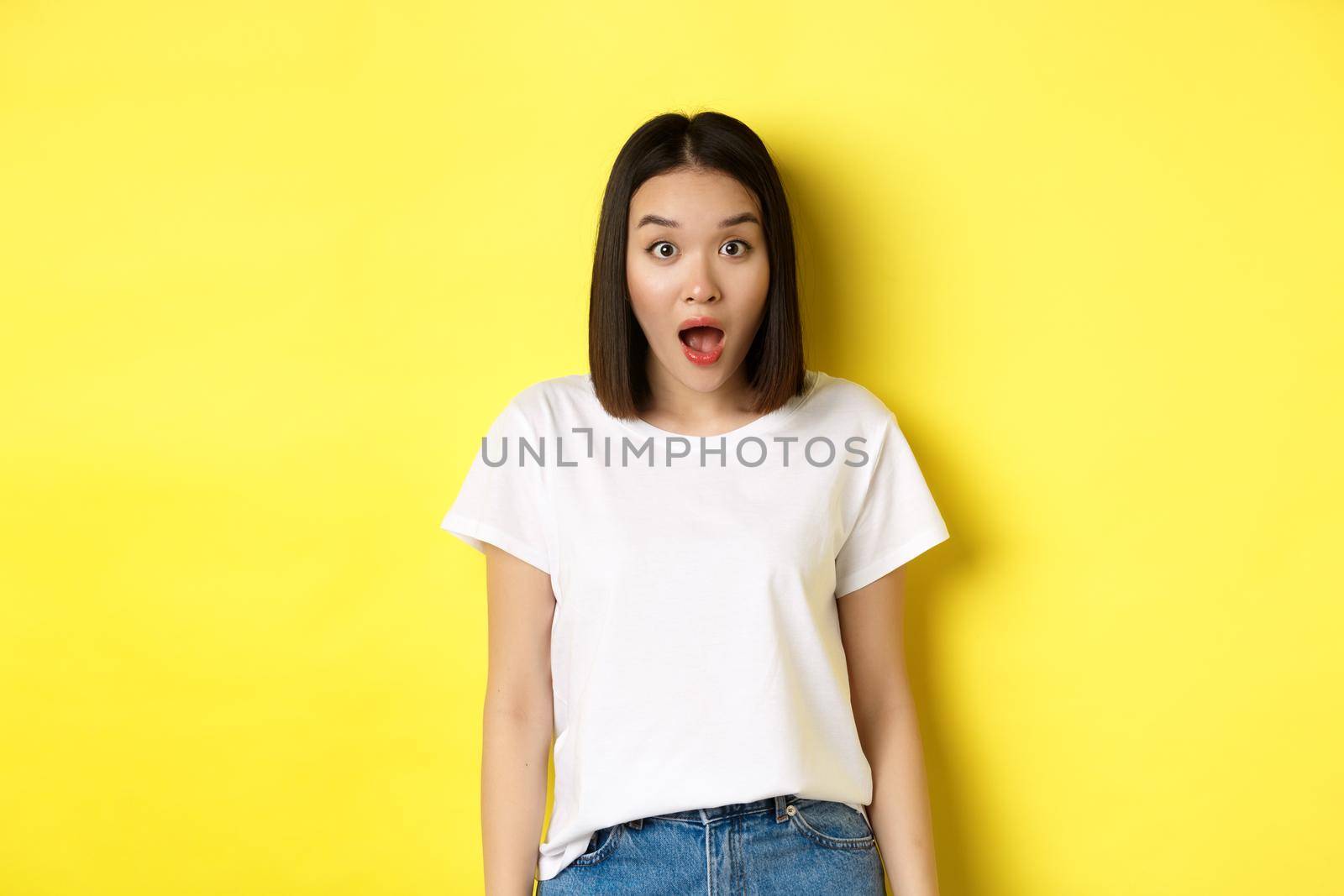 Beauty and fashion concept. Surprised asian girl open mouth and staring at camera, drop jaw impressed, standing over yellow background by Benzoix
