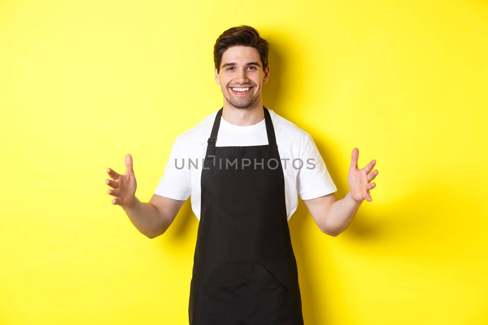 Smiling waiter in black apron holding your logo or box, spread hands as if carry something large, standing over yellow background by Benzoix