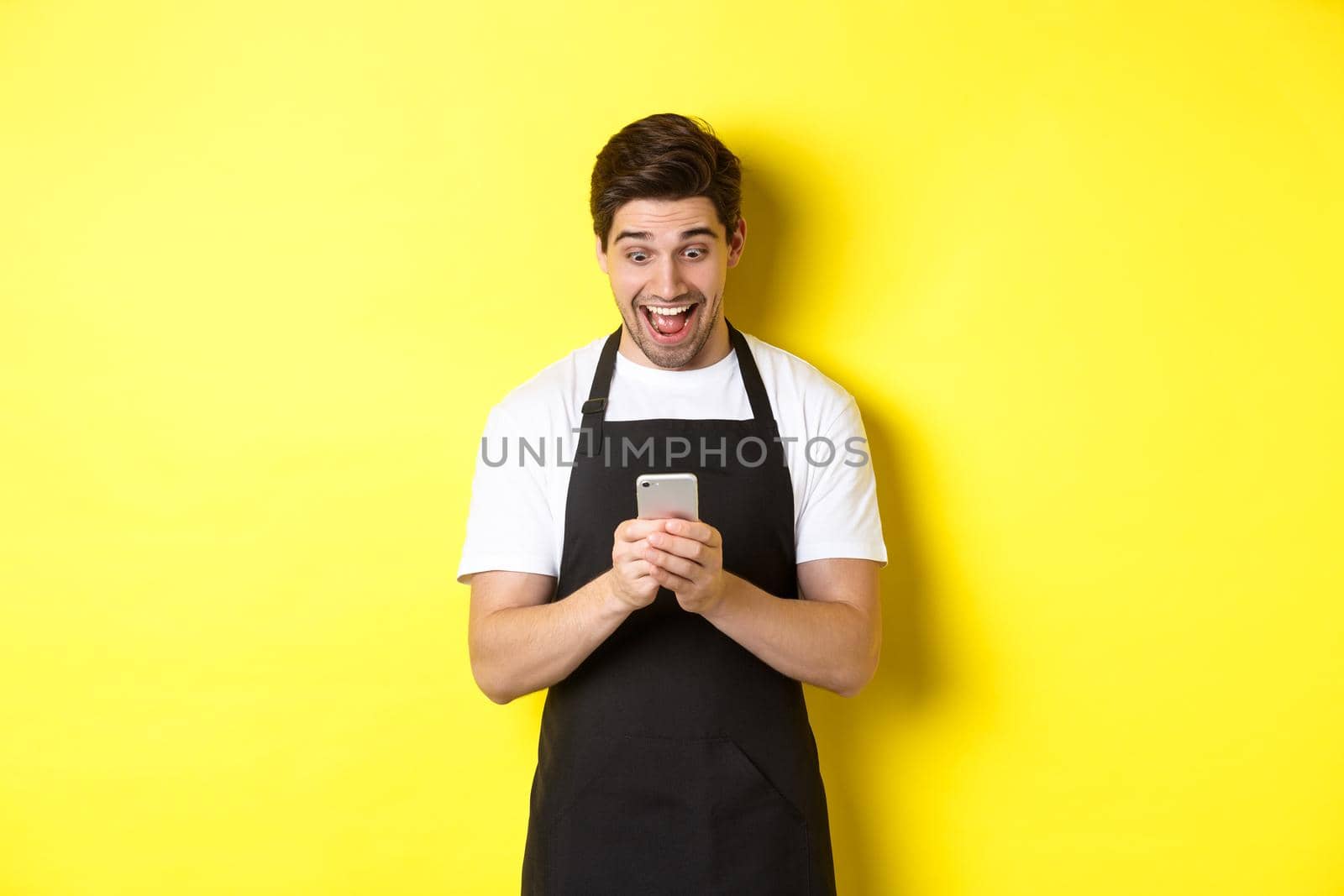 Barista looking surprised as reading message on mobile phone, standing in black apron against yellow background.