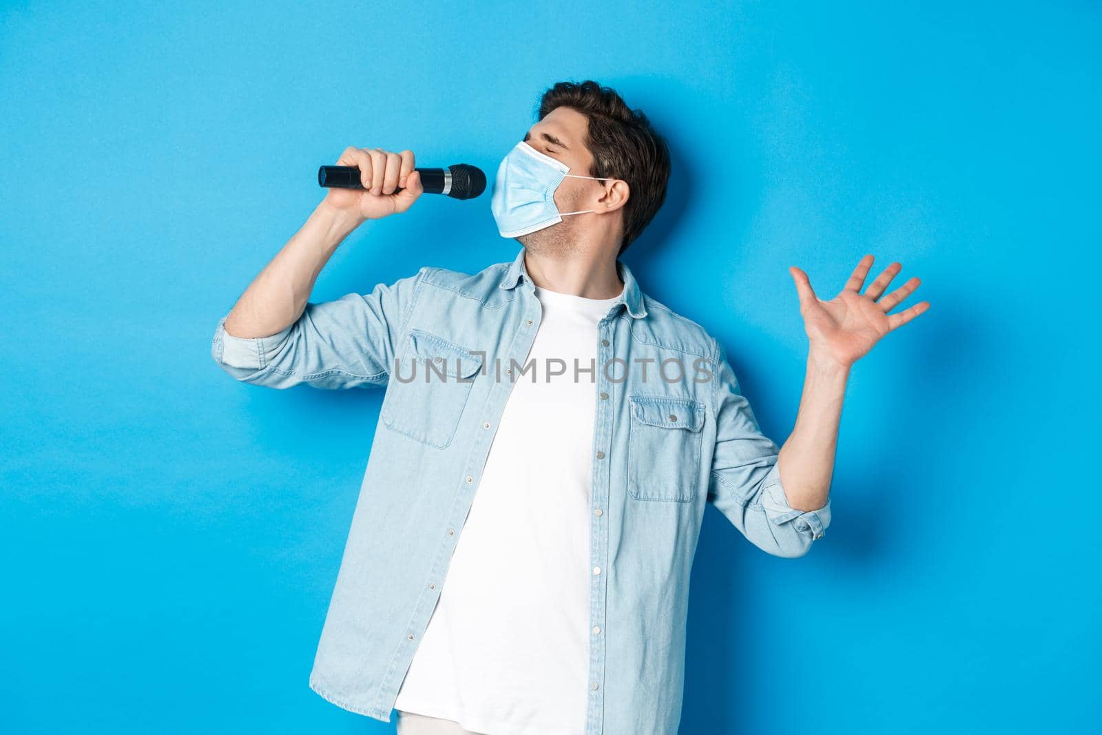 Handsome guy in medical mask singing in microphone, standing over blue background by Benzoix