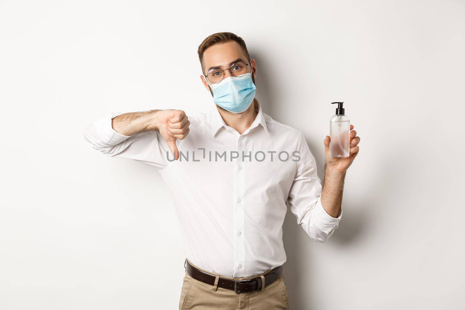 Covid-19, social distancing and quarantine concept. Office worker in medical mask displeased, showing hand sanitizer and thumb down, standing over white background by Benzoix