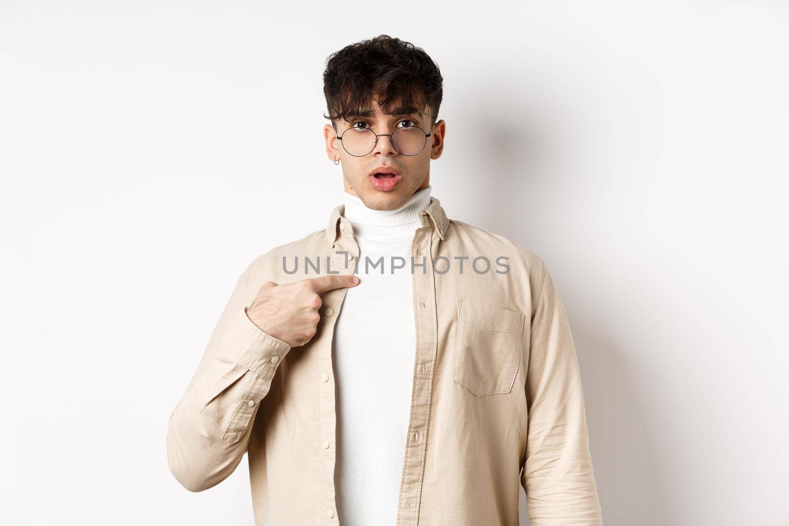 Confused and surprised young man pointing at himself, being chosen or accused, standing on white background.