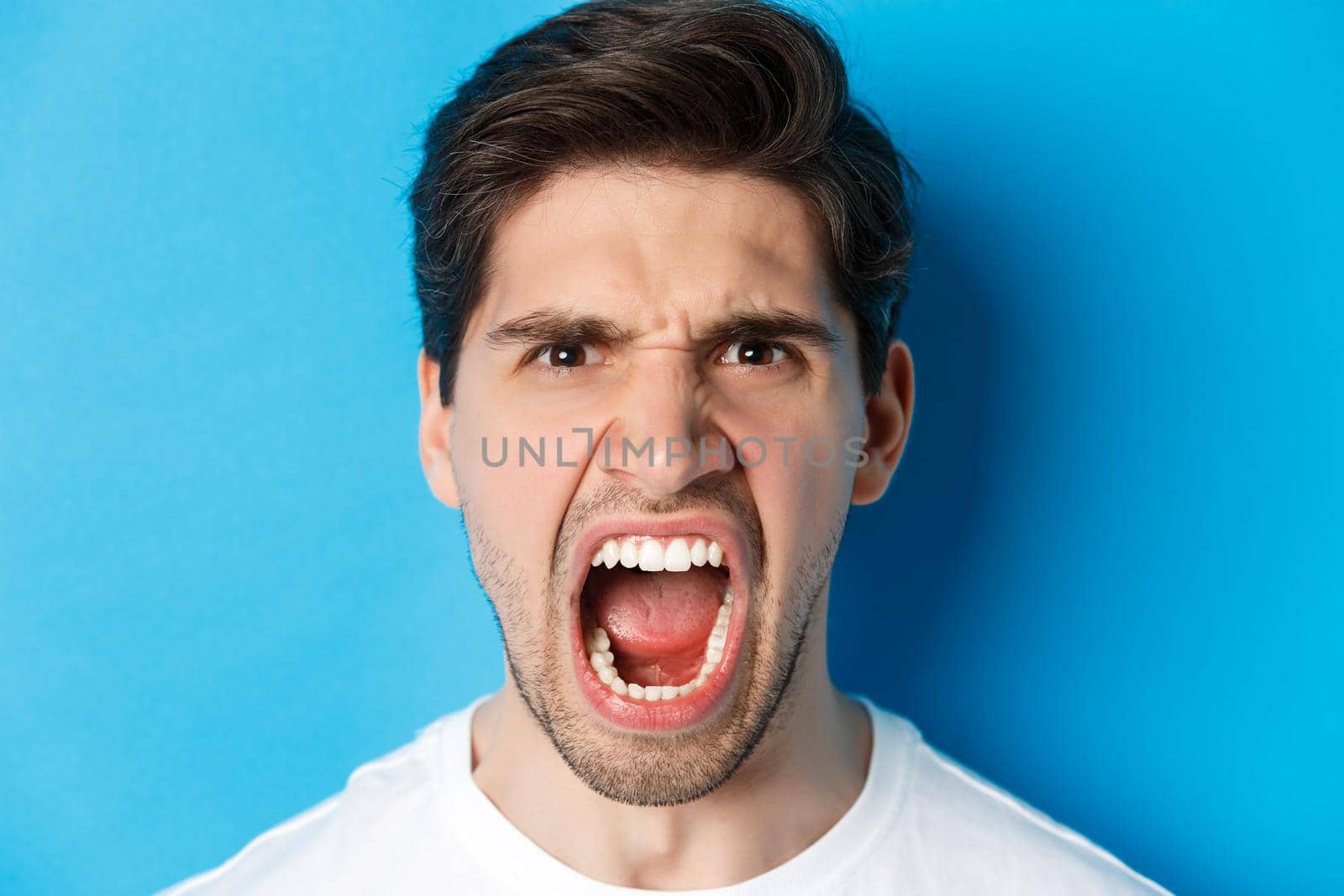 Head shot of angry man shouting and looking with hatred, standing mad against blue background by Benzoix