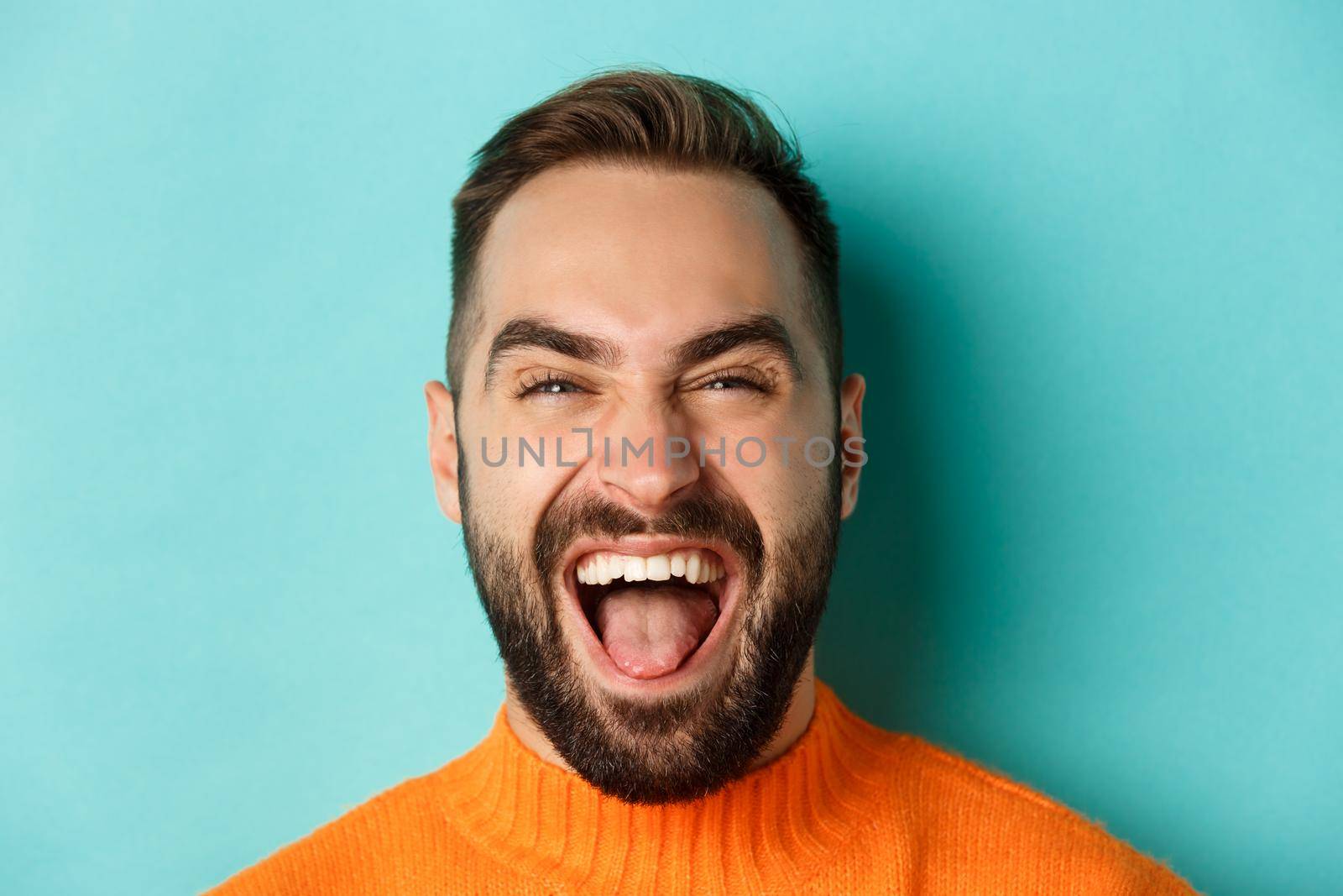 Headshot of happy and carefree man with beard, laughing and looking at camera, standing in orange sweater by Benzoix