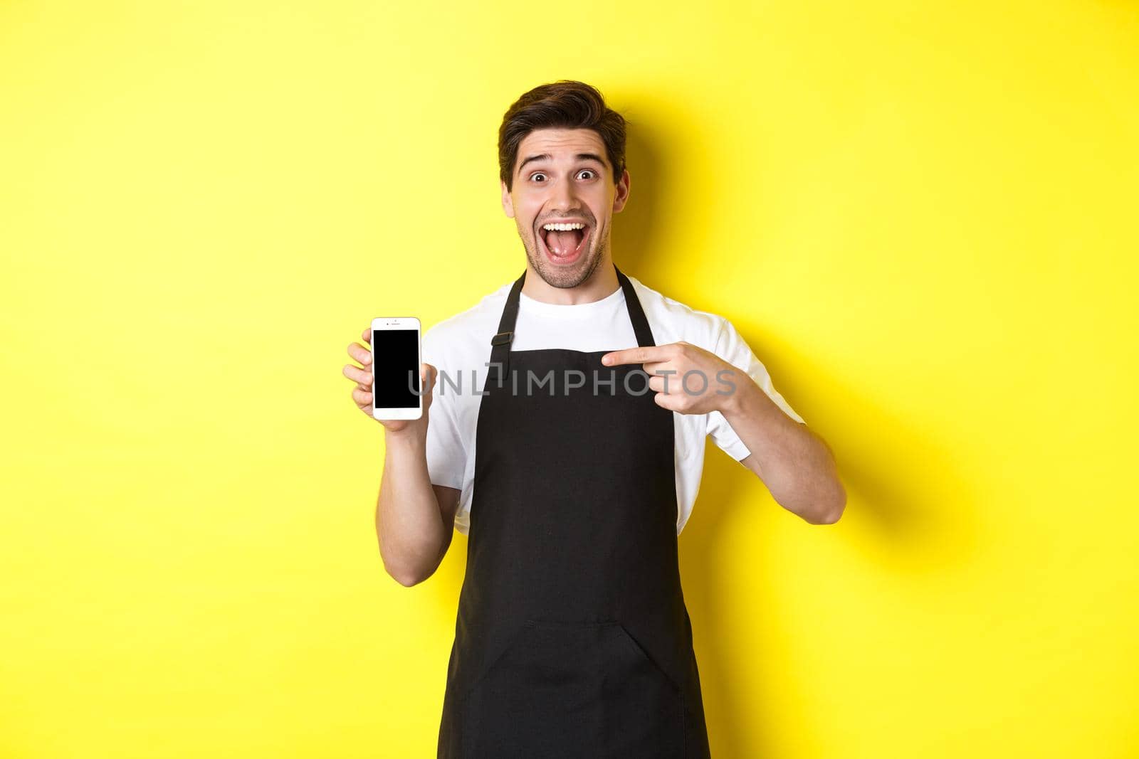 Handsome barista in black apron pointing finger at mobile screen, showing app and smiling, standing over yellow background.