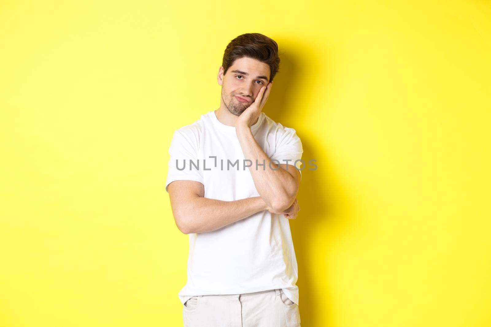 Bored and unamused man looking with indifference, standing in white clothes over yellow background.