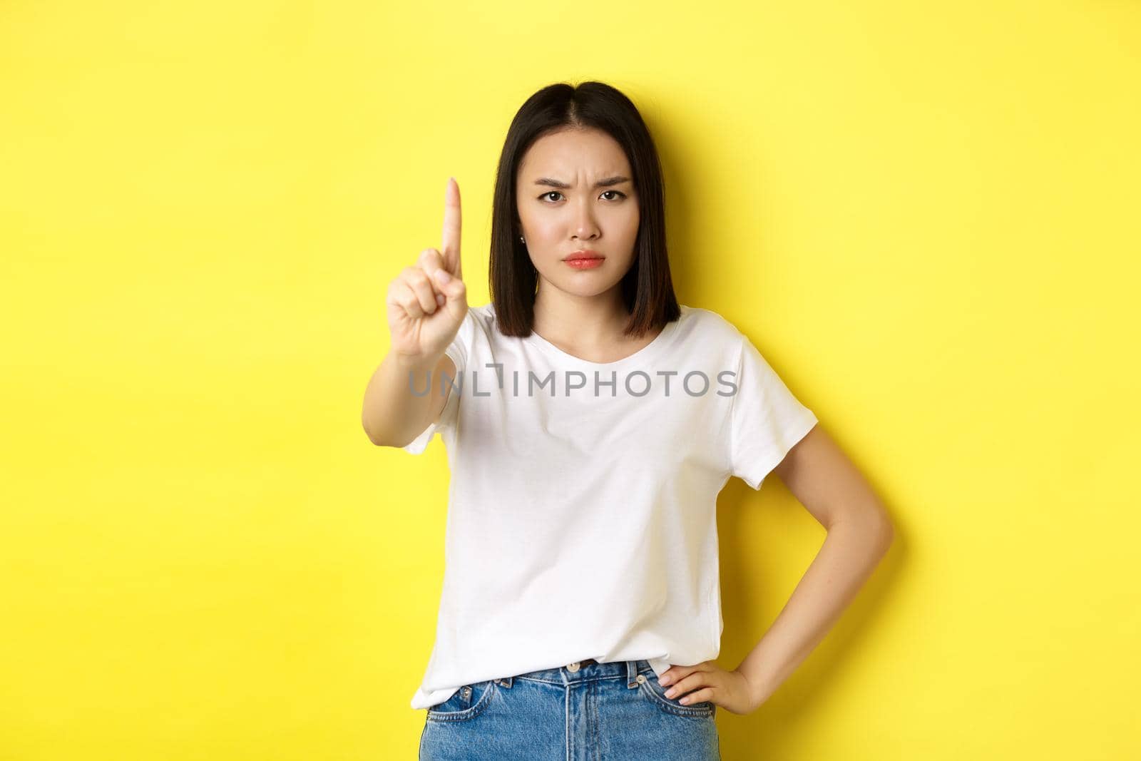 Confident and serious woman tell no, showing extended finger to stop and prohibit something bad, frowning and looking at camera self-assured, standing over yellow background.