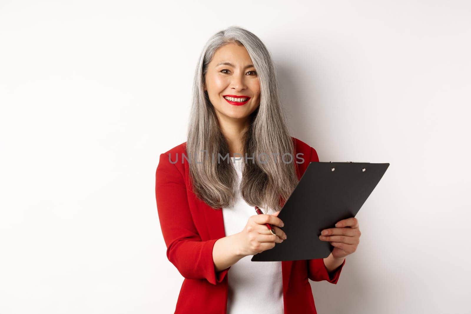 Elegant asian senior businesswoman working with documents, holding pen and clipboard, signing contract and smiling, standing over white background by Benzoix