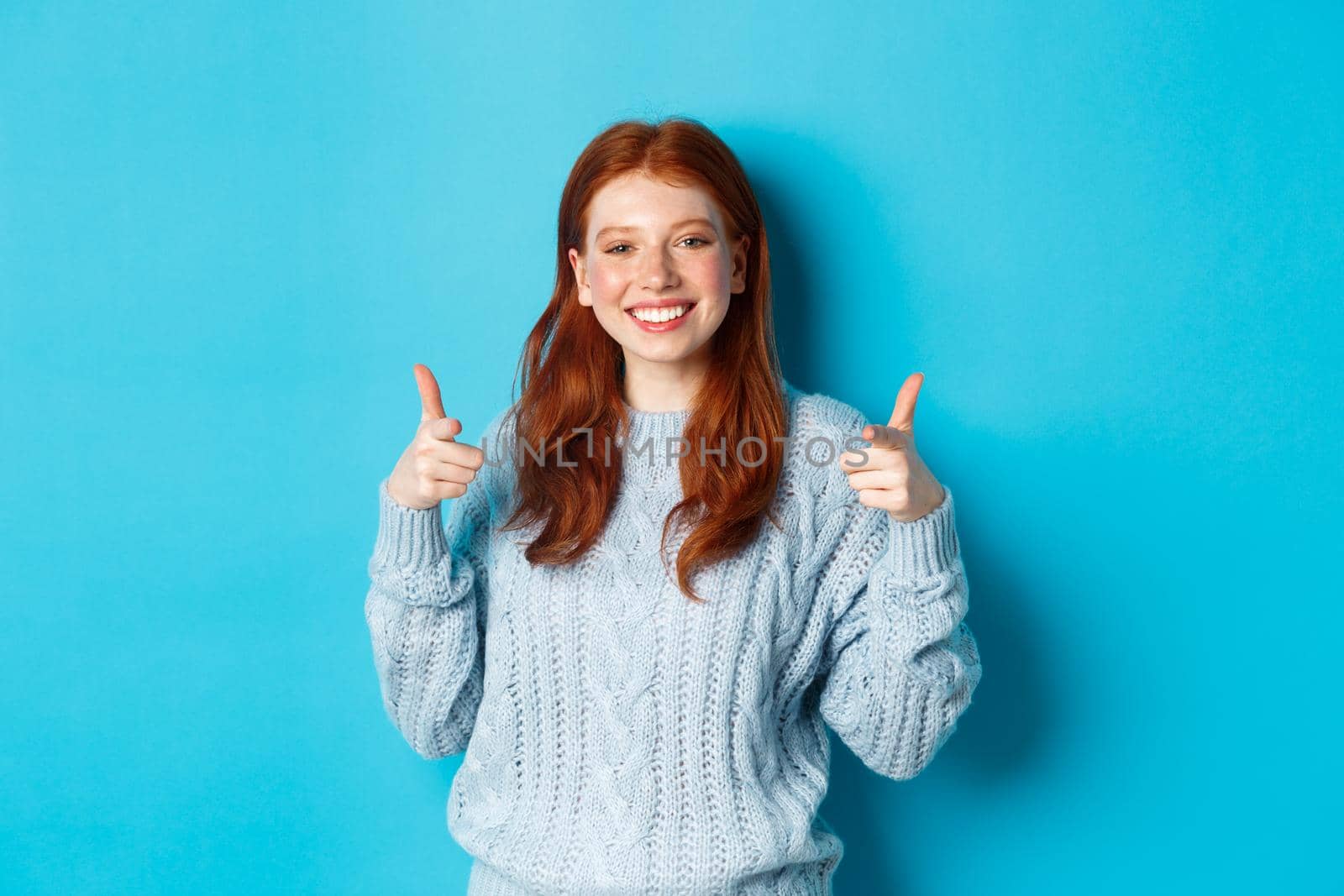 Cheerful teen girl with red hair, pointing fingers at camera and smiling, congratulating or praising you, standing over blue background.