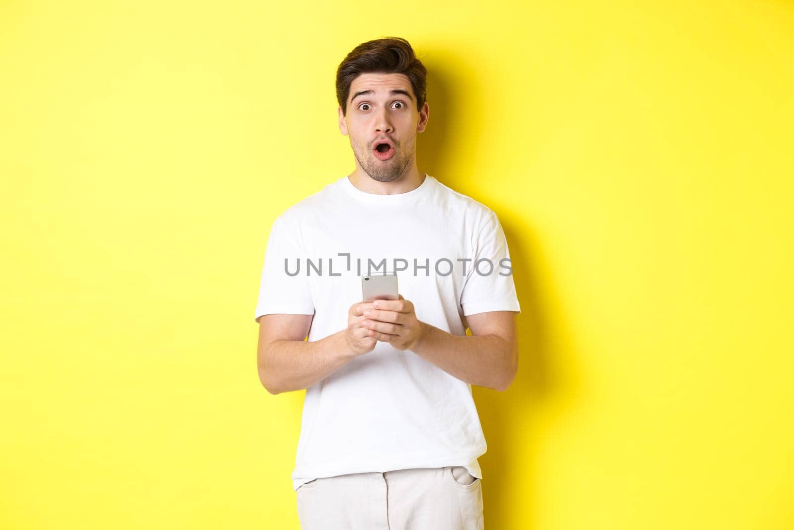 Man looking surprised, using smartphone, open mouth and saying wow, standing against yellow background. Copy space