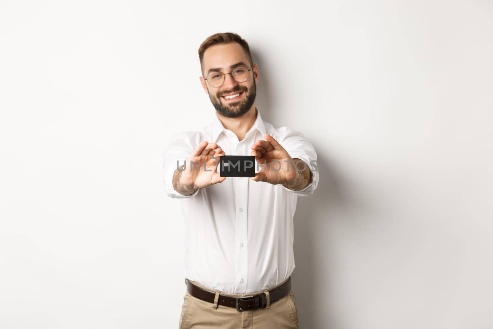 Handsome man in glasses holding a credit card, smiling pleased, standing over white background by Benzoix