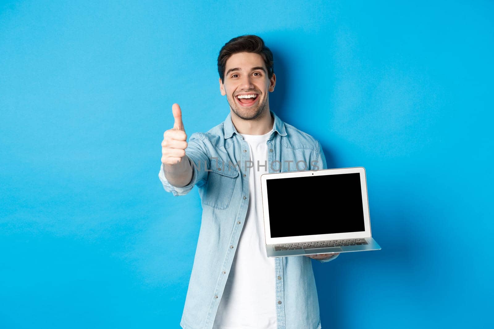 Image of satisfied handsome man showing laptop screen, thumbs-up in approval, like website or internet, standing over blue background.
