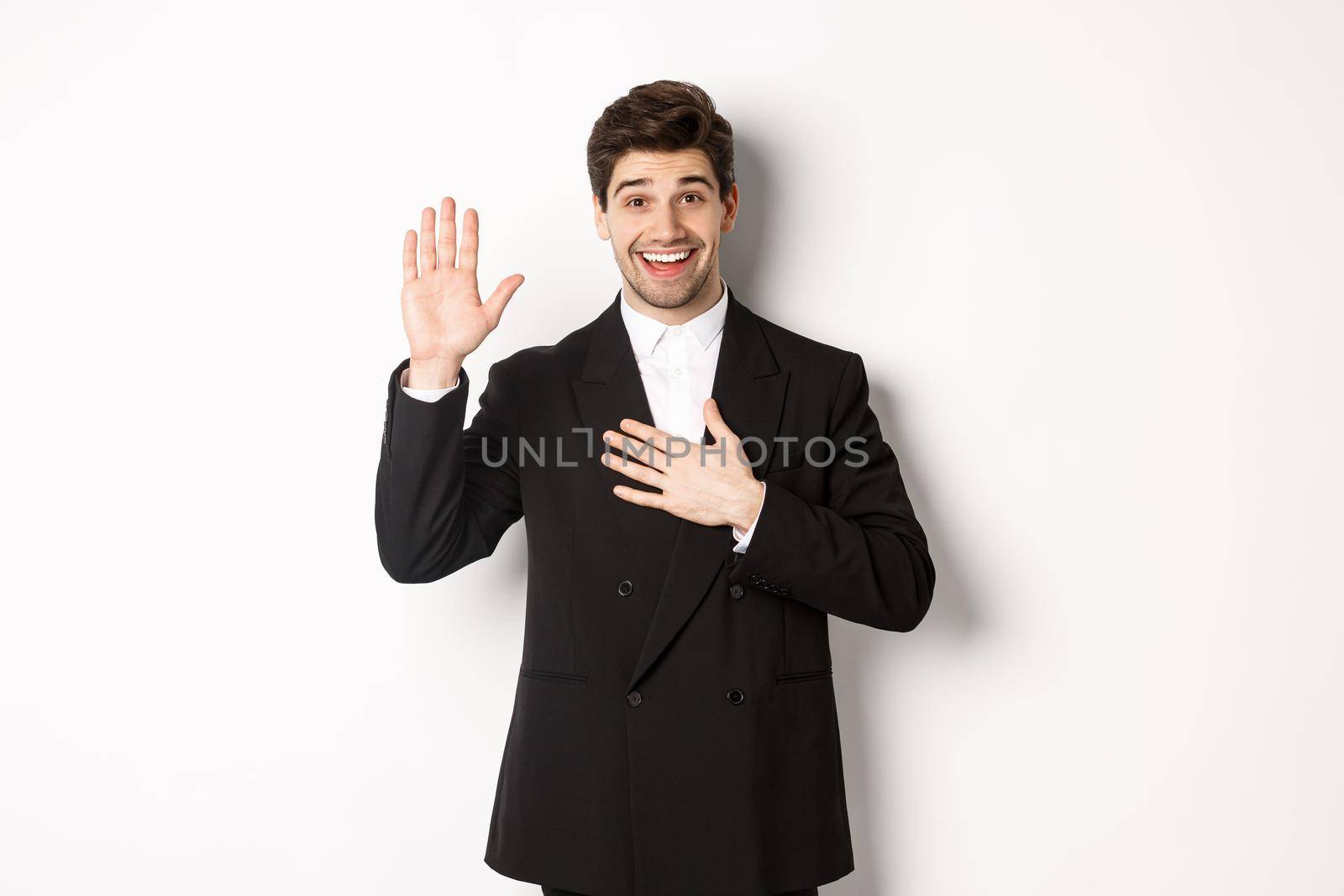 Image of handsome smiling guy telling the truth, raising on arm to promise something, standing over white background in black suit.