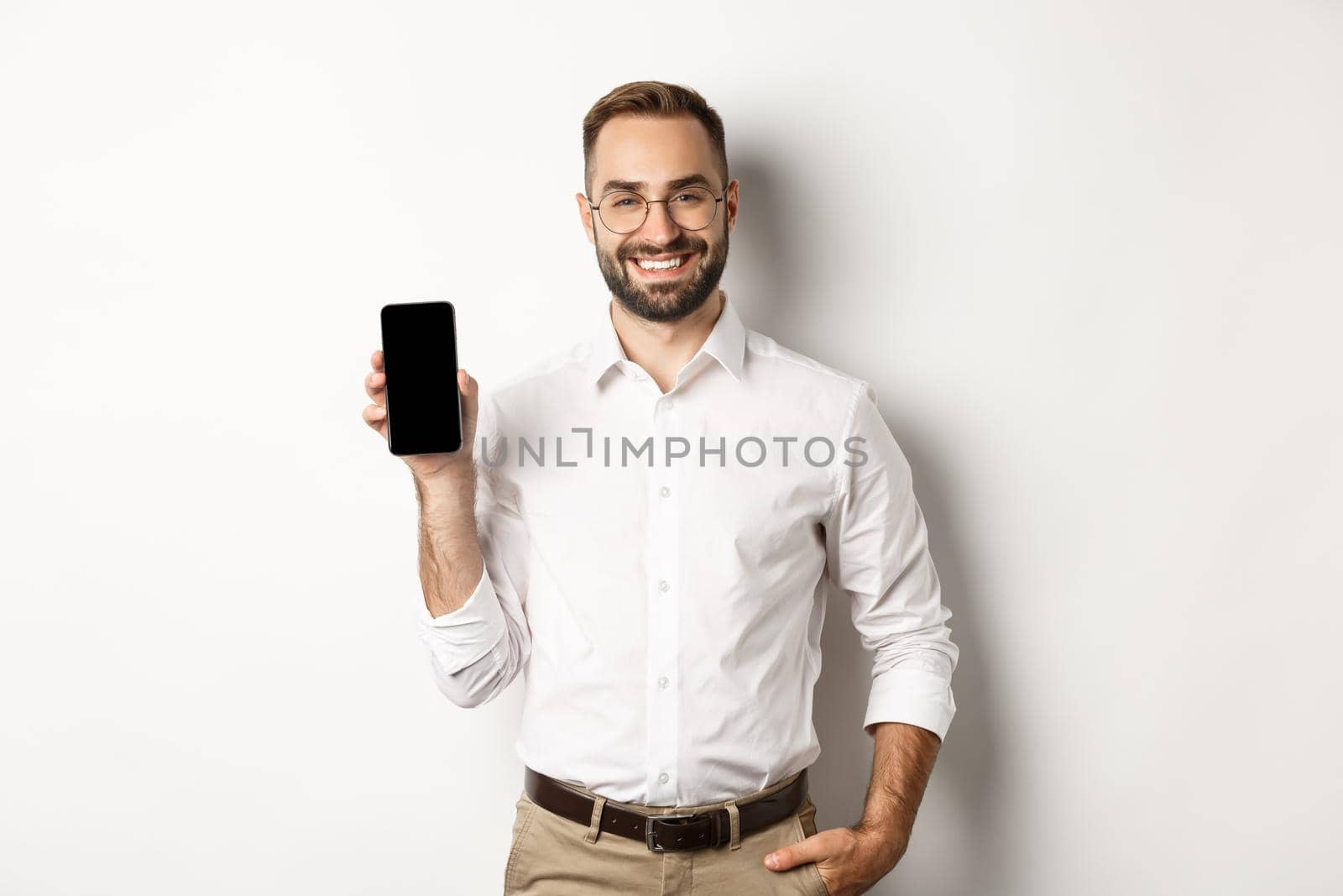 Satisfied business man showing mobile screen, smiling proudly, standing over white background.