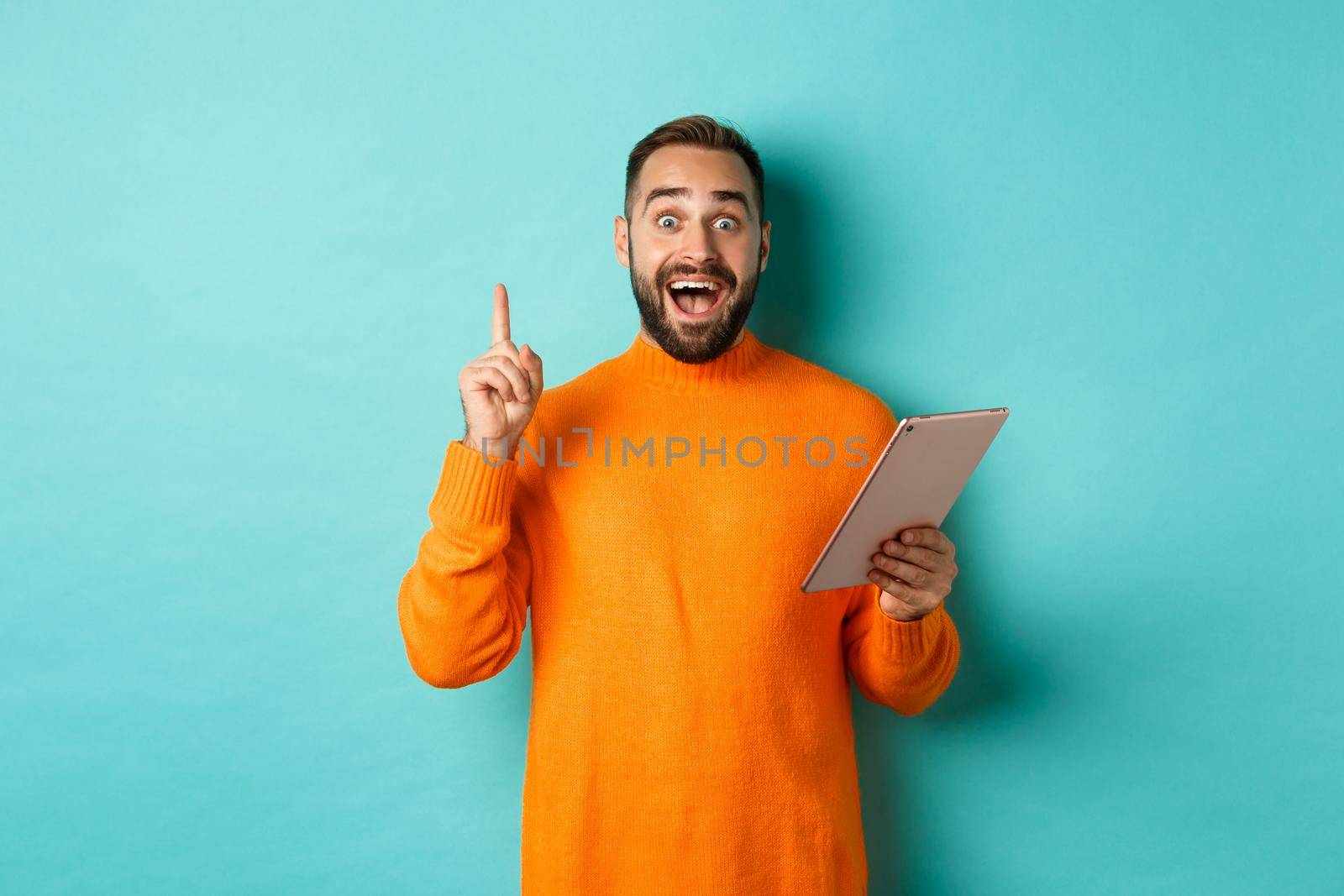 Excited adult man using digital tablet and having an idea, raising finger eureka sign, standing over light blue background.