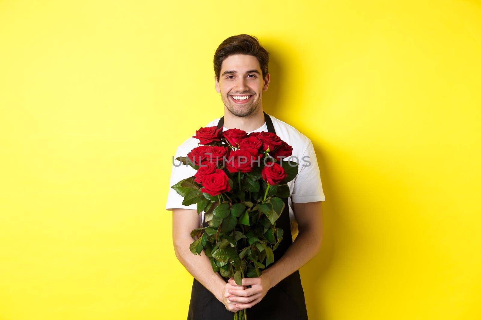 Smiling florist in black apron holding flowers, selling bouquet of roses, standing over yellow background.