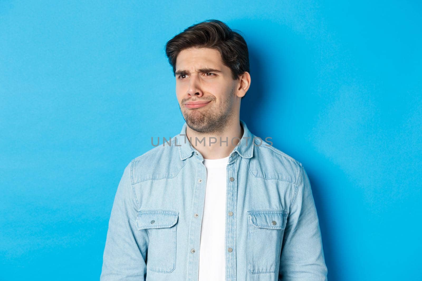 Close-up of upset and disappointed man grimacing, looking dissatisfied left, cringe from bad promo banner, standing over blue background by Benzoix