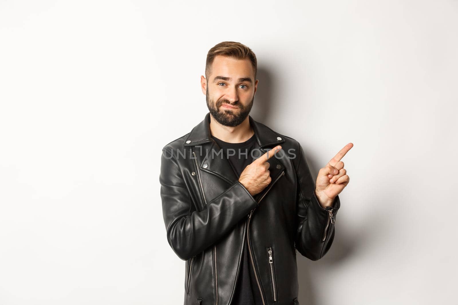 Skeptical and doubtful guy in black leather jacket, shrugging while pointing at upper right corner promo offer, standing over white background.