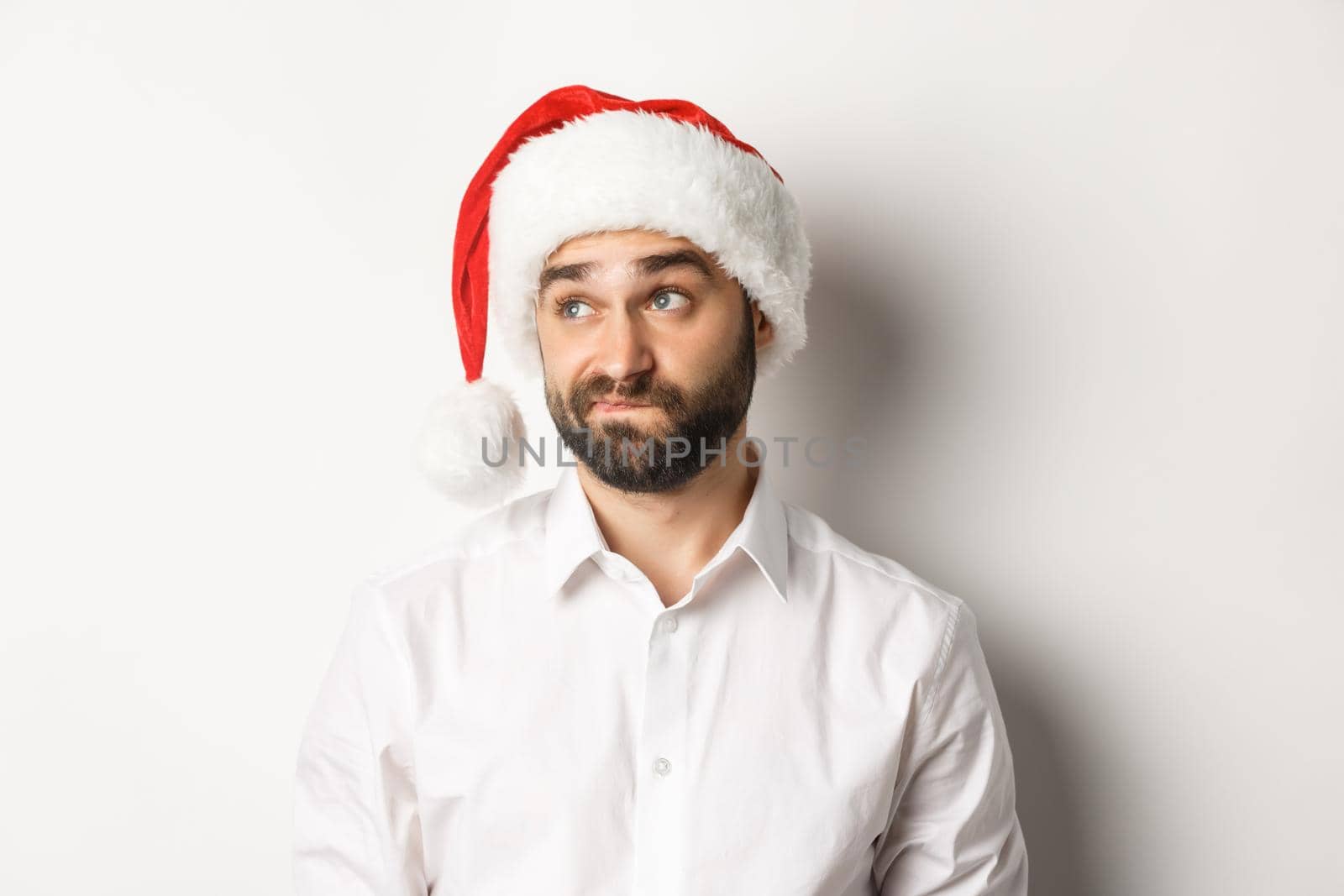 Close-up of skeptical guy in santa hat looking doubtful left, grimacing with hesitation, white background by Benzoix