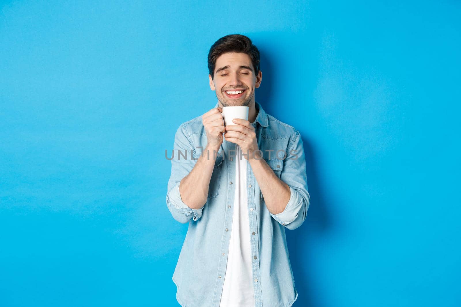 Satisfied man enjoying cup of tea or coffee, holding mug with pleased smile, standing against blue background.