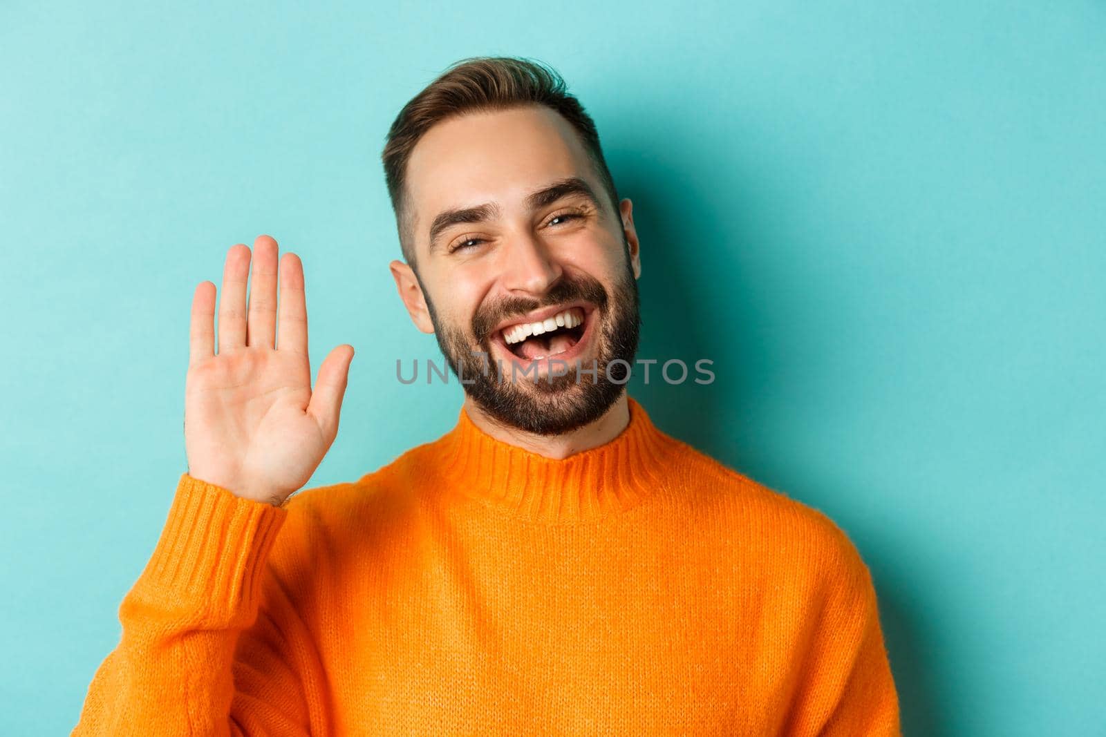 Close-up of friendly young man smiling, waving hand to say hello, greeting you, standing over light blue background.