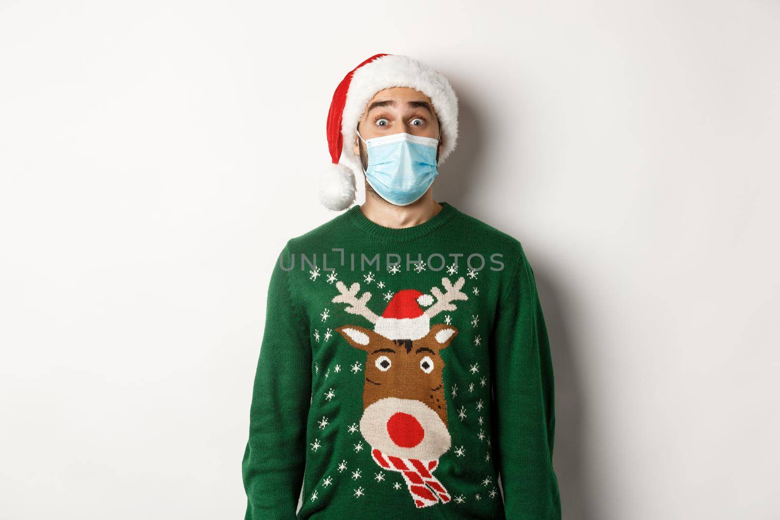 Christmas during pandemic, covid-19 concept. Surprised guy in medical mask, Santa hat and sweater celebrating New Year party, standing over white background.