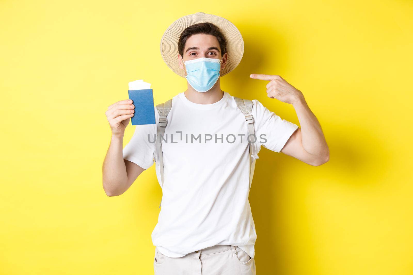 Concept of covid-19, tourism and pandemic. Man tourist showing passport, travelling in medical mask for protection from coronavirus, yellow background.
