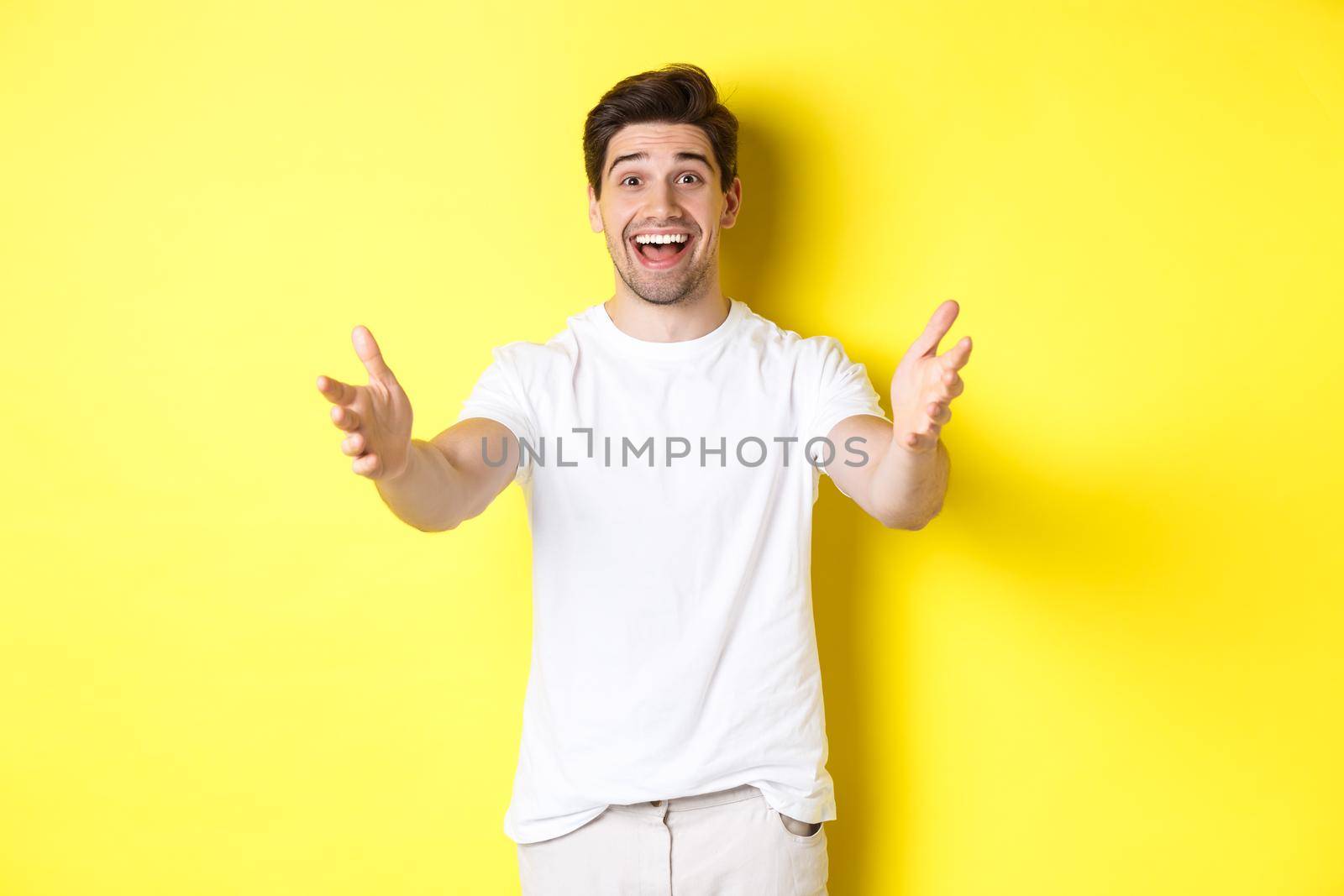 Excited handsome guy stretching hands forward, reaching for hug, receiving gift, standing over yellow background.