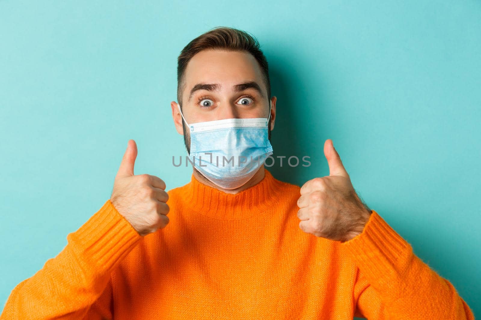 Covid-19, social distancing and quarantine concept. Close-up of satisfied male model in medical mask showing thumbs-up, praising and agreeing, standing over light blue background.