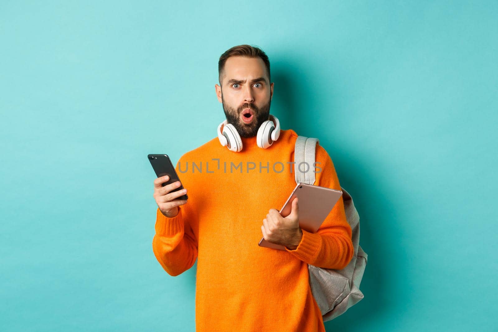 Handsome man student with headphones and backpack, holding digital tablet and smartphone, staring confused at camera, standing against turquoise background.