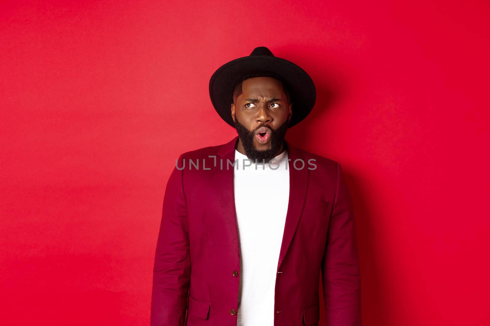 Fashion and party concept. Shocked and displeased black man staring at something unpleasant, looking upper left corner and frowning, standing against red background.