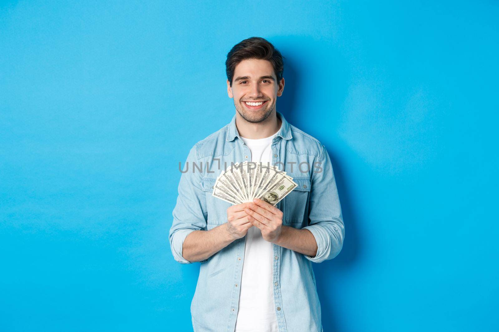 Handsome smiling man holding money, concept of finance and banking, standing over blue background by Benzoix