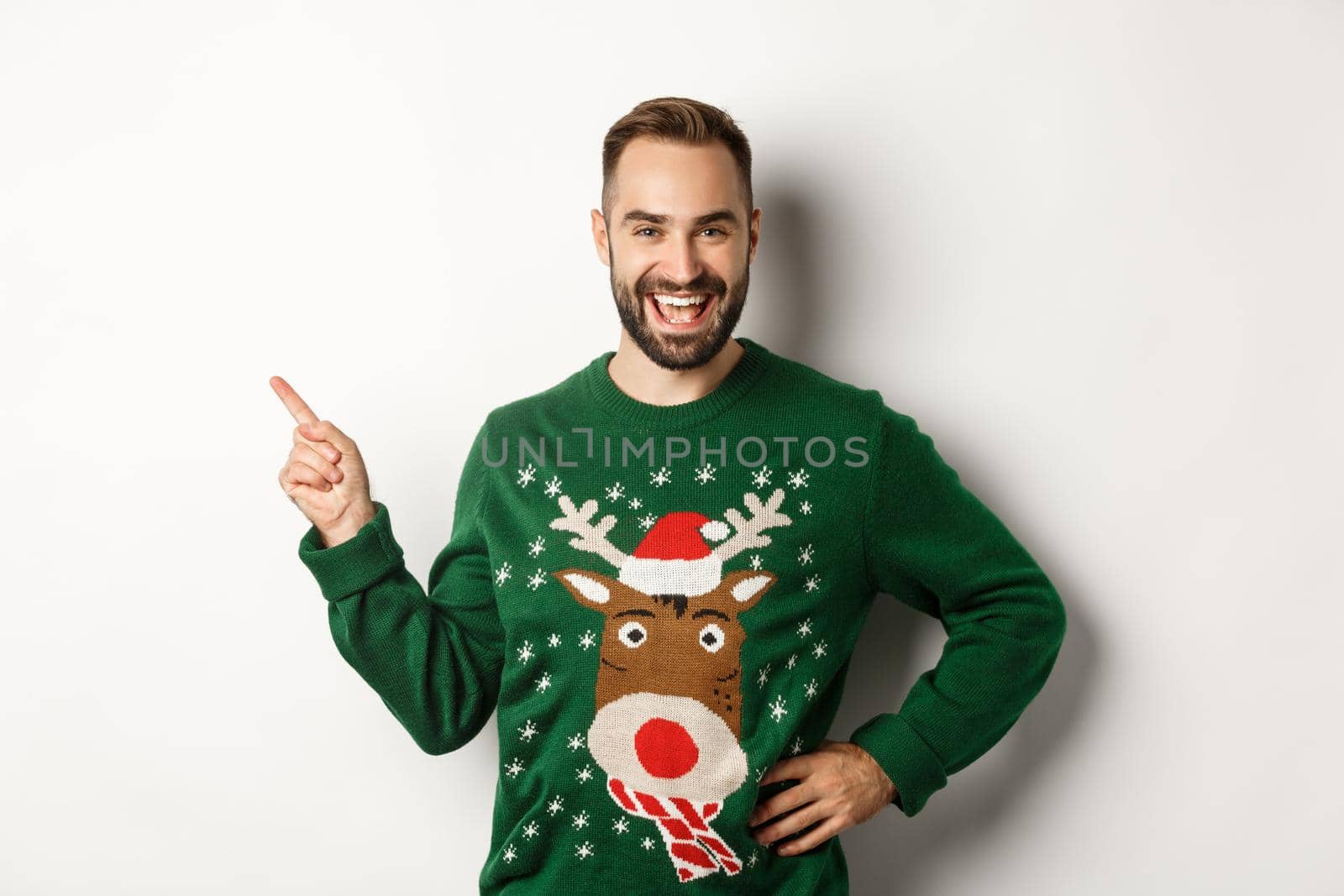 New year celebration and winter holidays concept. Handsome bearded male model in christmas sweater laughing, pointing finger at upper left corner logo, white background.