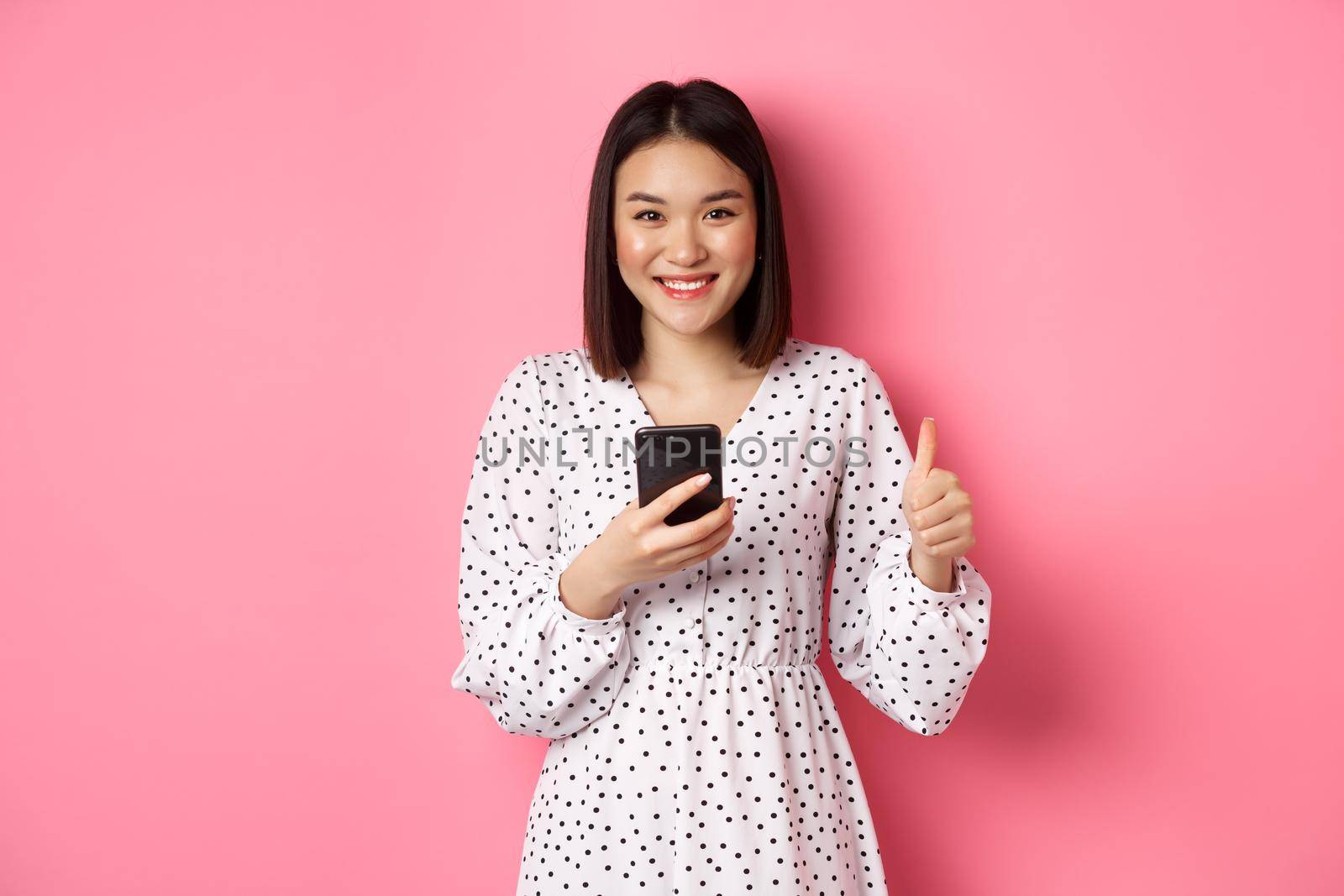 Online shopping and beauty concept. Satisfied asian female customer showing thumbs-up, making purchase in internet on smartphone, standing over pink background by Benzoix