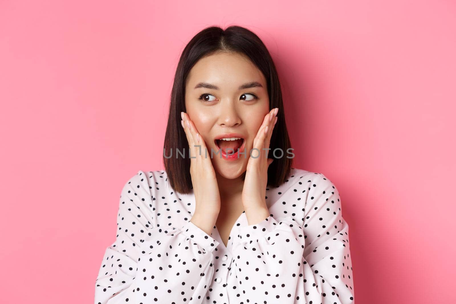 Close-up of beautiful asian woman looking surprised and excited, hear amazing news, looking left and rejoicing, standing against pink background.