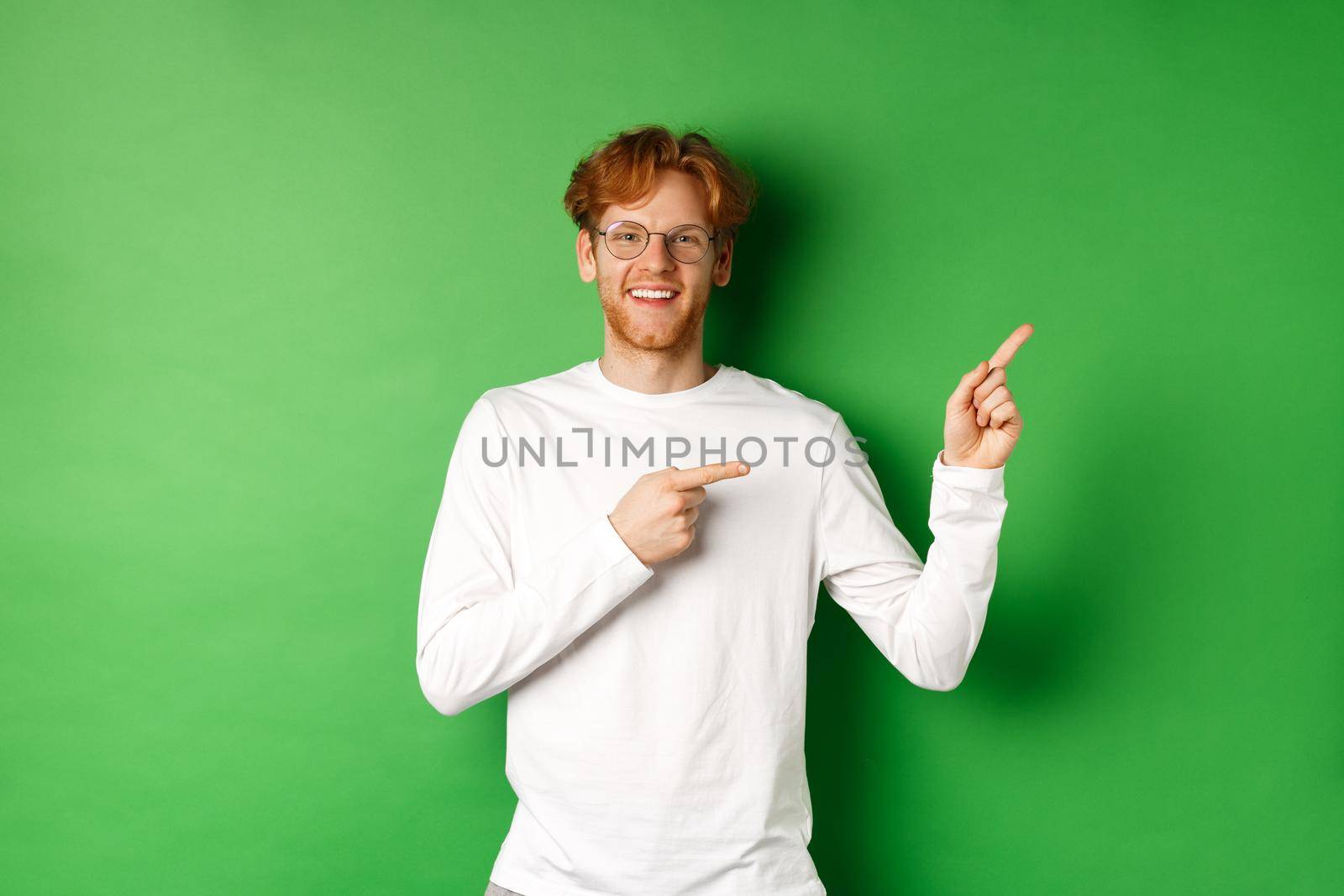 Handsome young man with ginger hair and glasses, pointing fingers right at copy space and smiling, standing over green background.