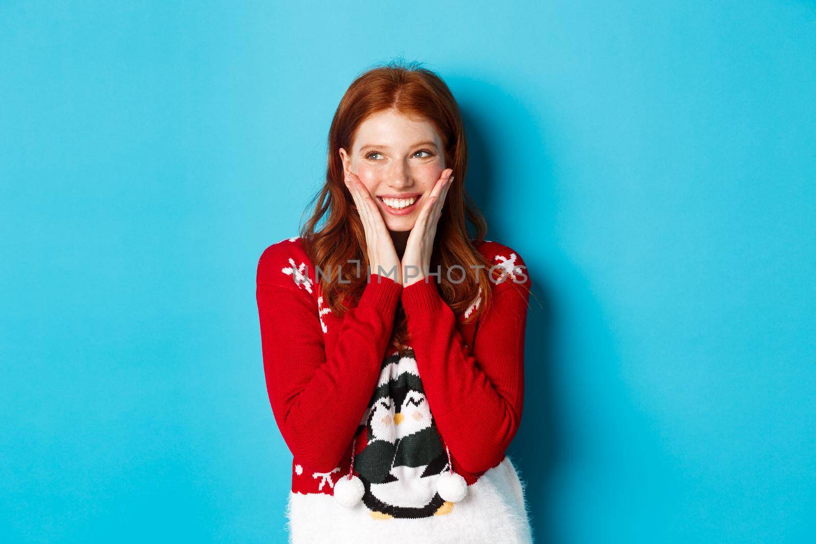 Winter holidays and Christmas Eve concept. Adorable redhead girl blushing and touching cheeks from happiness, smiling and looking at upper left corner promo, blue background by Benzoix