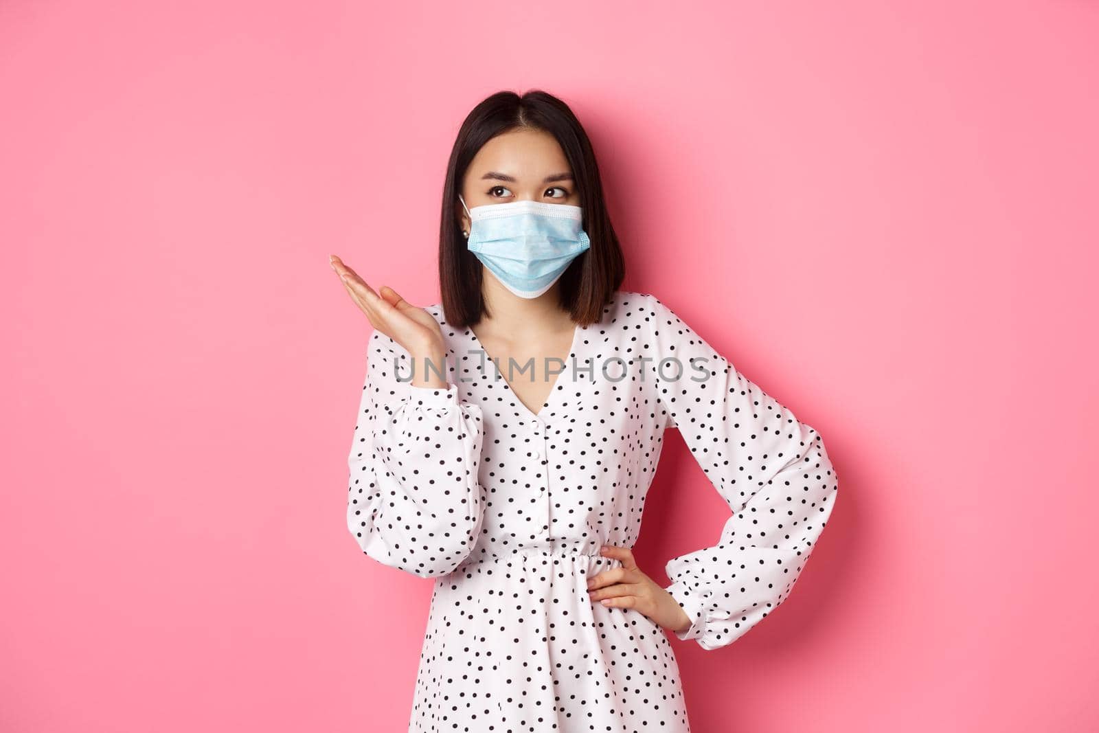 Covid-19, quarantine and lifestyle concept. Lovely asian woman in face mask raising hand, standing in dress over pink background.